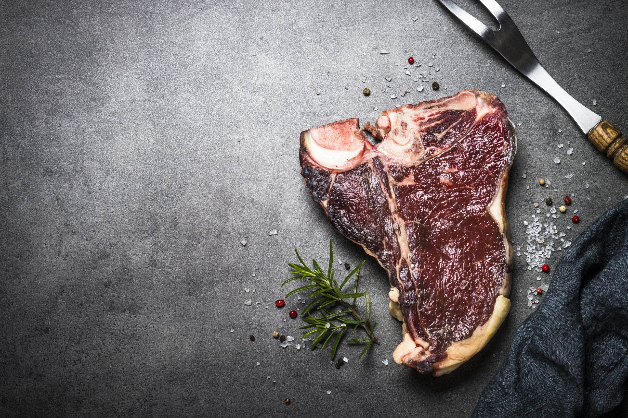 Rohes Steak mit Knochen auf einer dunklen Fläche, garniert mit Rosmarin und grobem Salz
